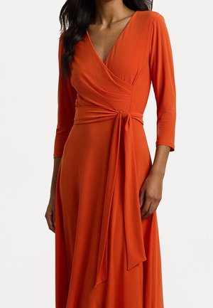 Kobieta ubrana w długą, głęboko pomarańczową sukienkę kopertową z paskiem zawiązanym w talii, dekoltem w serek i spódnicą o kroju A na jednolitym białym tle.