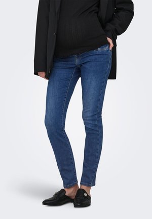 Person iført sort jakke, sort top, blå skinny jeans og sorte slip-on loafers, stående med den ene hånd i lommen.
