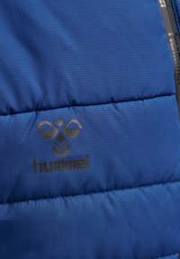 Chaqueta azul hinchada con textura de cuadrícula, que presenta un logo negro de "hummel". La tela es suave y el diseño incluye costuras horizontales.