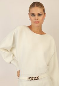 Weißer gerippter Pullover mit oversized Silhouette, breitem Halsausschnitt und metallischer Kettenakzent an der Taille. Weiche Textur und lässige Passform.