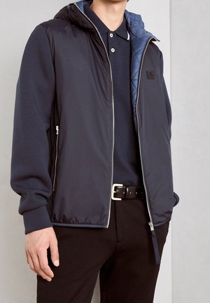 Mann trägt eine marineblaue Kapuzenweste mit Reißverschluss über einem marineblauen Poloshirt, schwarze Hose und schwarzen Gürtel mit goldener Schnalle, steht vor einer grauen Wand.