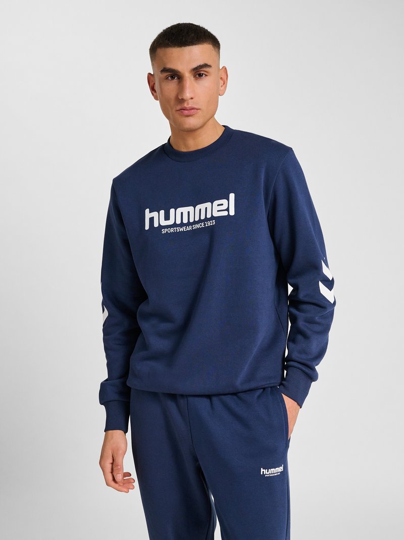 Mann trägt navyblaue Hummel-Sweatshirt und passende Sweatpants mit weißem Logo-Schriftzug und Chevron-Designs auf den Ärmeln.