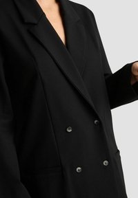 Czarny dwurzędowy żakiet noszony przez osobę z widoczną częściową szyją i ręką, na jednolitym jasnoszarym tle.