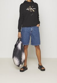 Svart huvtröja med "Calvin Klein Jeans"-logotyp, blå denimshorts, svarta plattformsandaler och en tie-dye-handväska.