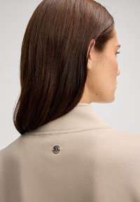 Blazer beige con una texture liscia, con un accento circolare in logo argento sul retro. Design sartoriale con spalle strutturate.