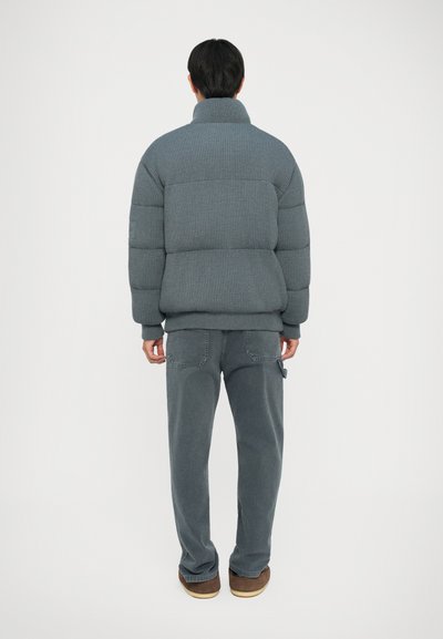 Siva pufasta jakna s rebrastom teksturom i visokim ovratnikom, uparen s odgovarajućim sivim hlačama. Outfit se odlikuje opuštenim krojem i minimalističkim dizajnom.