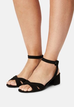 Pieds portant des sandales à talons blocs en daim noir avec des brides de cheville et des bandes croisées sur les orteils, sur fond blanc.