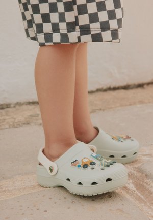 Kind trägt hellblaue Clogs, verziert mit Spielzeugauto- und Eieranhängern, steht auf Steinpflaster, mit sichtbaren karierte Shorts.