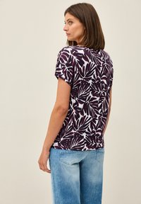 T-shirt à manches courtes en blanc avec un motif de plante violet audacieux. Matériau doux, coupe décontractée, encolure arrondie et manches roulées.