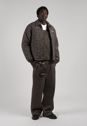 Un homme porte une veste marron texturée, un pantalon ample foncé, des chaussures noires et une casquette accrochée à sa ceinture, sur un fond de studio gris.