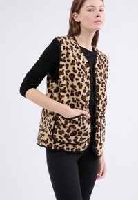 Kamizelka polarowa w lamparcim wzorze z czarnymi wykończeniami, z okrągłym dekoltem i dwiema przednimi kieszeniami, noszona na czarną bluzkę z długim rękawem.