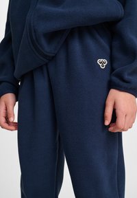 Marinblå fleece-träningsoverall med en dragkedjeficka, elastiskt midjeband och en liten broderad logotyp på byxorna. Mjuk textur.