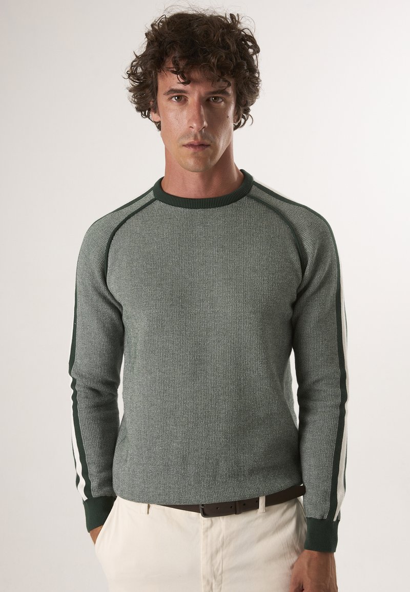 Groene gebreide sweater met een gestructureerd patroon, met contrasterende groene en witte strepen langs de mouwen en een geribde halslijn.