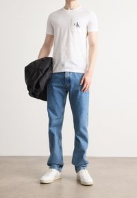 Muž oblečený v bílém tričku Calvin Klein, modrých džínách a bílých teniskách, drží černou bundu na jednobarevném pozadí.