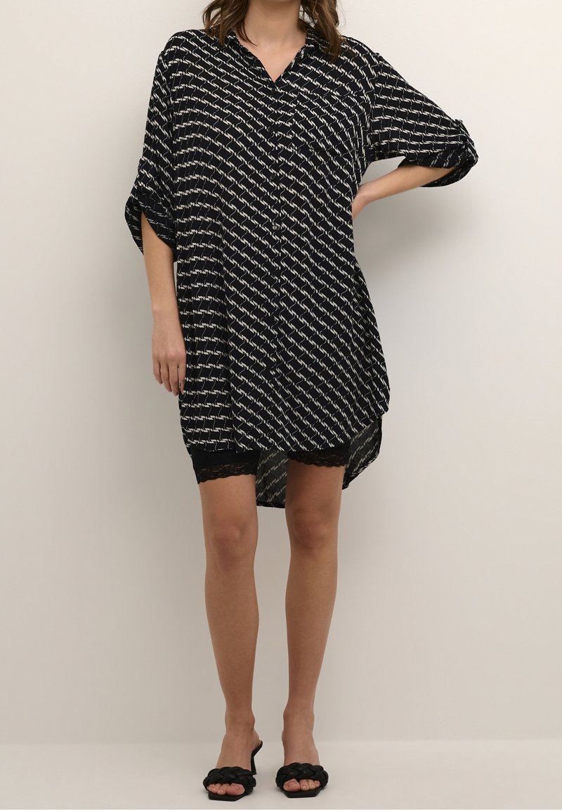 Femme portant une robe chemise ample à motifs noir et blanc, avec des manches retroussées et des sandales slides noires tressées.