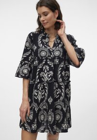 Marinblå klänning med vita blommönster, flera lager design och trumpetärmar. Funktioner inkluderar en knappförsedd placket och mjukt, strukturerat tyg.