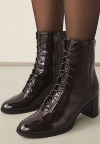 Brune patentlæder-ankelstøvler med firkantet tå, snøring foran og lynlås på siden. De har en chunky hæl og en glat tekstur.