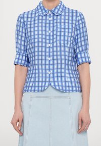 Camicia a quadri blu con colletto arrotondato, maniche corte e tasca frontale. Realizzata in un tessuto liscio con una leggera lucentezza.