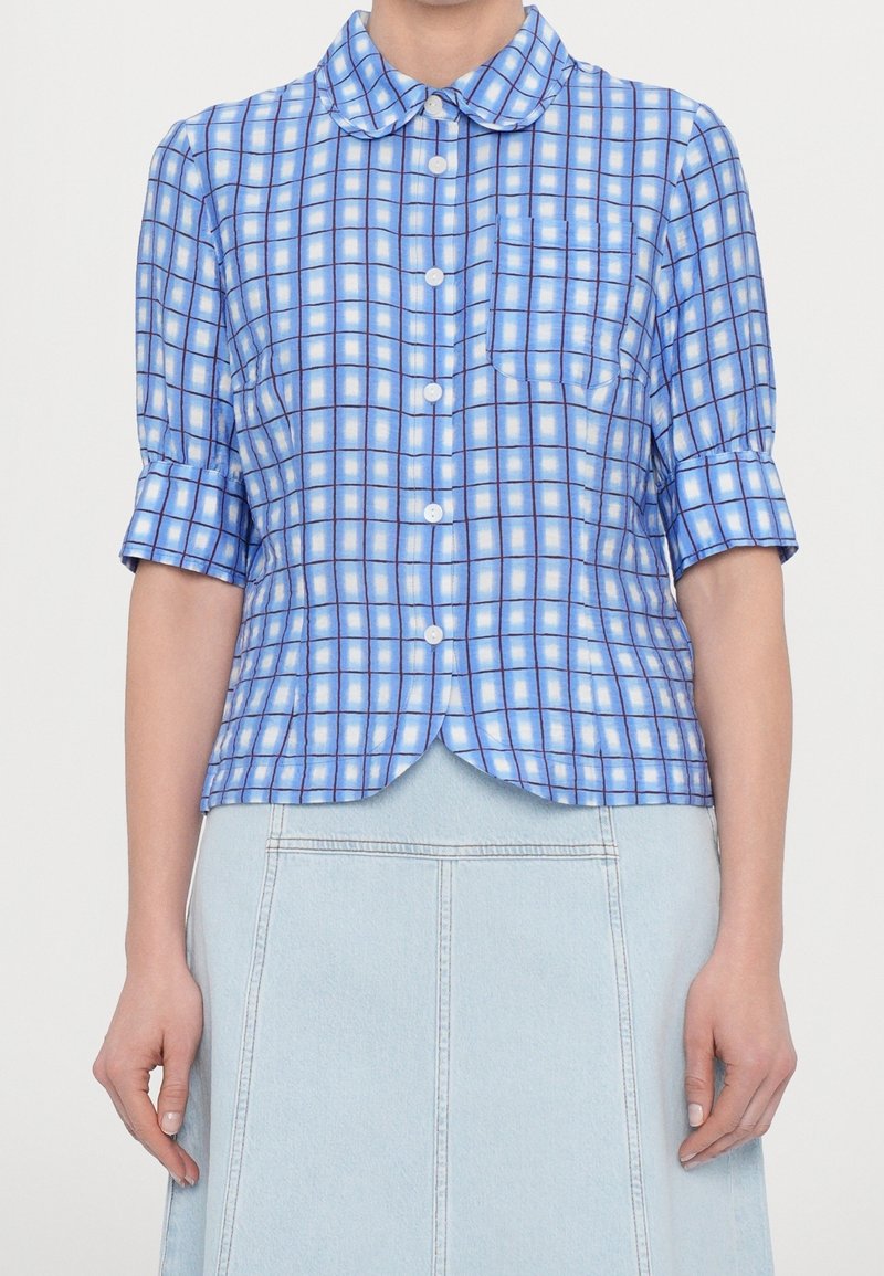 Camicia a quadri blu con colletto arrotondato, maniche corte e tasca frontale. Realizzata in un tessuto liscio con una leggera lucentezza.