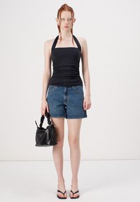 Zwarte haltertop, blauwe denim shorts en zwarte handtas. Model in zwarte teenslippers staat tegen een effen witte achtergrond.