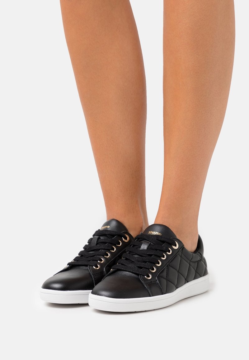 Baskets en cuir matelassé noir avec semelles blanches portées sur des jambes nues sur un fond clair uni.