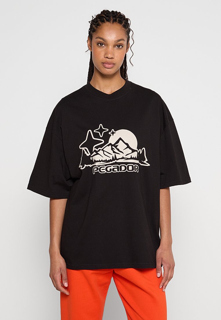 T-shirt surdimensionné noir en coton, avec un design graphique de montagnes et d'étoiles en beige clair, accompagné du texte "PEGADOR" en bas.