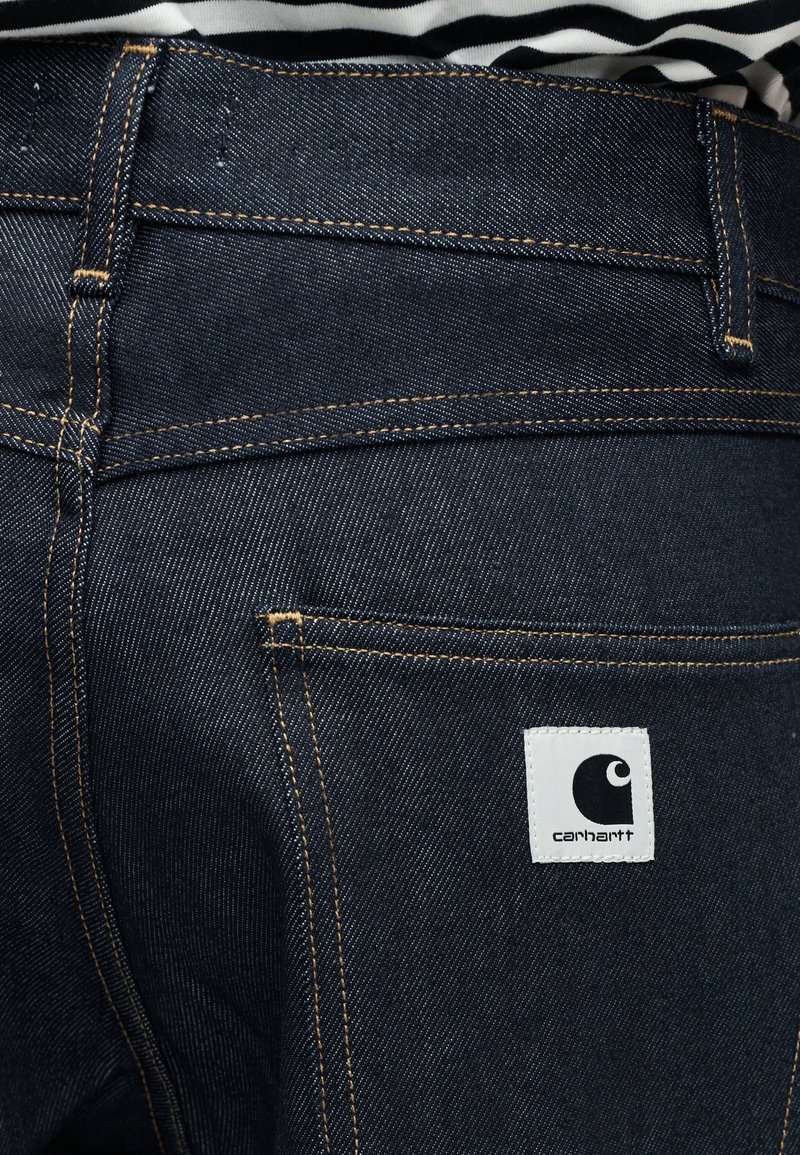 Mørkeblå denimjeans med orange syninger og et hvidt Carhartt logo patch på baglommen, båret med en stribet skjorte.