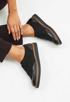 Scarpe in suede nere con accenti neri lucidi, design con lacci, suola piatta e bordi dettagliati. Indossate con pantaloni scuri su uno sfondo chiaro.