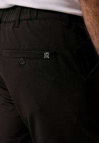 Zwarte broek van gladde stof, met een achterzak met knop en een klein geborduurd logo. Eenvoudig, tailormade ontwerp.