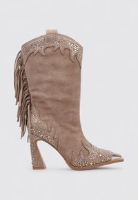 Bota vaquera de ante beige con punta afilada, puntera metálica, intrincados patrones recortados, flecos a los lados y un tacón ancho.