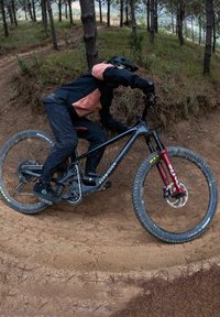 Horský bicykl s černým rámem a červenými předními vidlicemi, umístěný na nerovném terénu. Má silné pneumatiky, řetěz a ochranu na řetězové vzpěře.