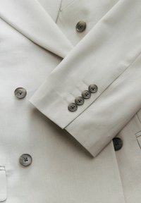 Blazer beige con tessuto testurizzato, design doppiopetto e cinque bottoni scuri sul polsino, con dettagli di cuciture accentuate.
