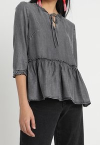 Šedý peplum top vyrobený z lehkého denimu, s kulatým výstřihem a tkaničkou, třičtvrtinovými rukávy a elastickými detaily.