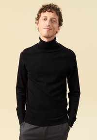 Schwarzer Rollkragenpullover mit langen Ärmeln, gerippten Bündchen und Saum. Weiche Textur, taillierte Passform. Getragen mit grauen Hosen.