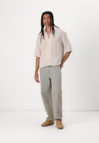 Helles beiges Kurzarm-Hemd mit Knopfleiste und Karomuster, kombiniert mit lockeren hellblauen Jeans und beigen Sneakers auf einem einfarbigen Hintergrund.