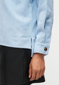 Γαλάζιο ριμπ τοπ με μακρύ μανίκι και μανσέτα με καφέ κουμπί, συνδυασμένο με μαύρη φούστα, και ένα χέρι που ακουμπάει δίπλα στο σώμα.