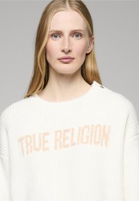 Maglione bianco lavorato a maglia con trama a coste e scritta "TRUE RELIGION" color pesca chiaro sul petto; presenta un bottone decorativo sulla spalla.