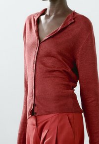 Red cardigan with a button-up front, ribbed hem, and a relaxed fit, paired with matching red trousers. Soft, knitted texture.