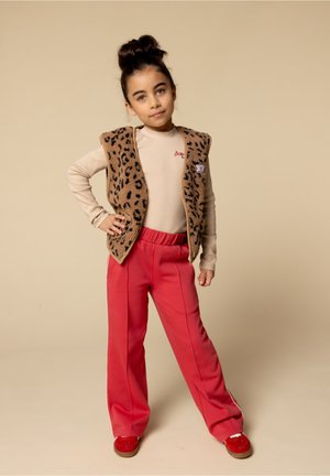 Meisje met donker haar in een bun, gekleed in een beige langemouwen shirt, een panterprint vest, rode broek en rode schoenen, staand met haar hand op haar heup.