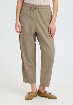 Pantalon beige à cordon, fuselé, porté avec des mocassins Mary Jane en daim beige, montré sur une personne debout sur un fond uni.