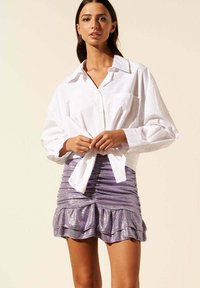 Chemise blanche à boutons portée avec une mini-jupe violette métallique comportant des couches à volants et une silhouette ajustée.