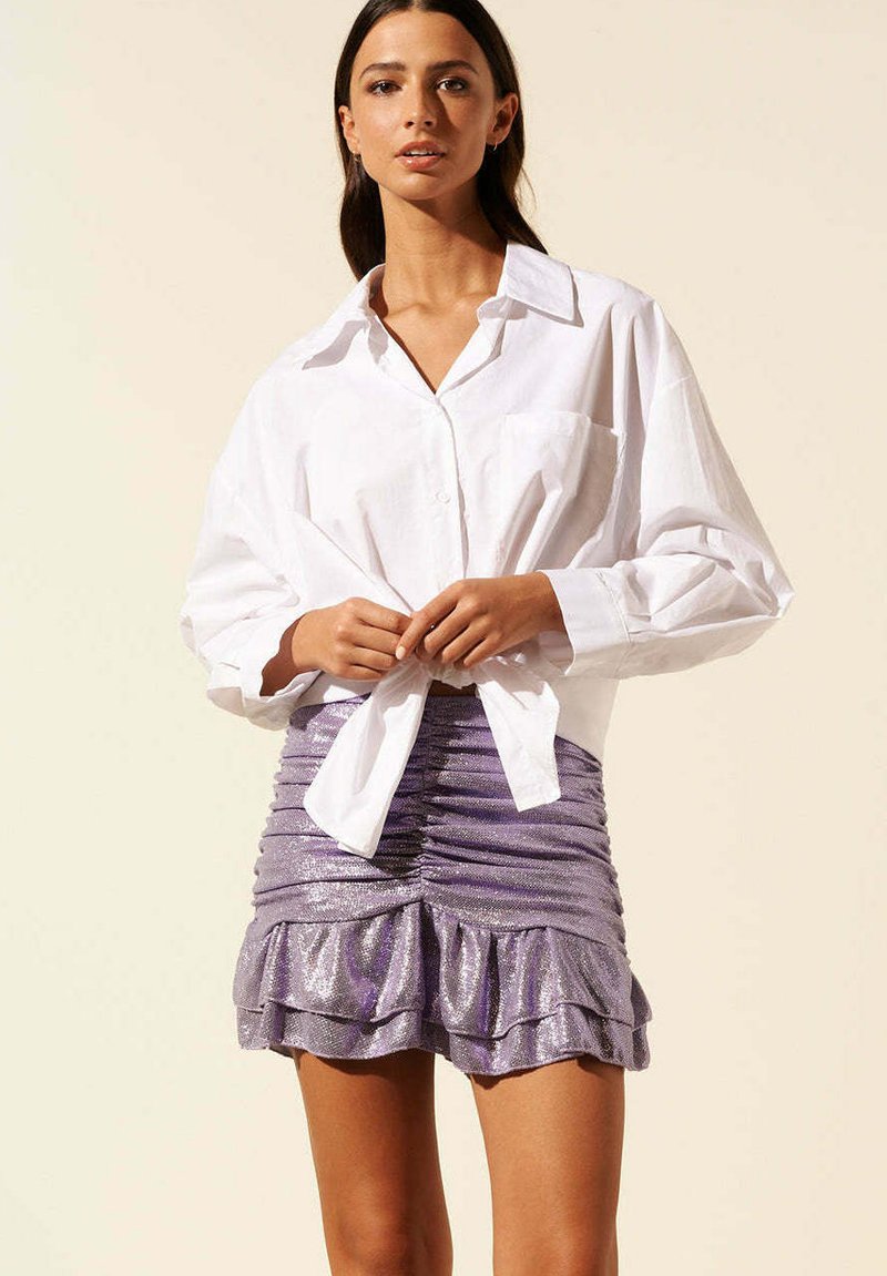 Chemise blanche à boutons portée avec une mini-jupe violette métallique comportant des couches à volants et une silhouette ajustée.