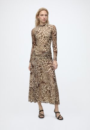 Femme portant une robe à manches longues, imprimé léopard marron et noir, avec un col montant et des sandales noires à talons hauts, debout devant un fond blanc.