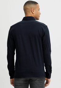 Hombre con cabello corto, vistiendo una camisa de manga larga de color azul marino oscuro y jeans grises, de pie de espaldas contra un fondo ligero y liso.