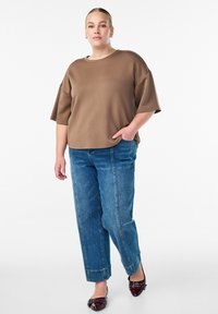 Pruun üleöö lõikega lühikeste varrukatega topp, paaritatud helesiniste kitsaste teksade ja burdeosvärvi madalate kingadega, näidates lõdva sobivust ja sujuvat tekstuuri.