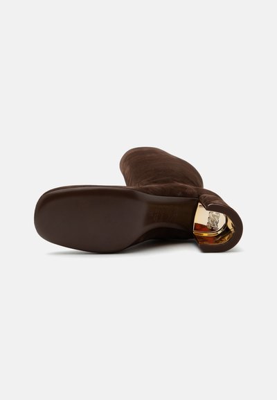 Botte en daim marron montant jusqu'au genou avec une semelle plate et un accent métallique doré à l'intérieur du talon courbé, vue du dessous.