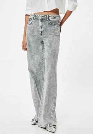 Jeans acampanados de talle alto en gris claro, con una textura desgastada y descolorida. Presentan cinco bolsillos y trabillas para el cinturón, combinados con zapatos metálicos de punta afilada.