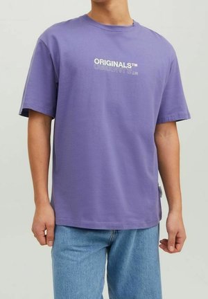 Homme portant un T-shirt violet avec le texte « ORIGINALS™ » et un jean bleu clair, debout devant un fond clair uni.