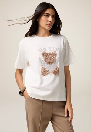T-shirt bianco a maniche corte con una grafica di un orso marrone peloso e il testo "NEW YORK". Disponibile con pantaloni beige e un braccialetto decorativo.