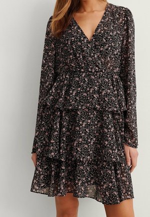 Kobieta ubranа w czarną kwiecistą sukienkę kopertową z długimi rękawami i falbanami na plisowanej spódnicy, stojąca na tle jasnej, gładkiej ściany.